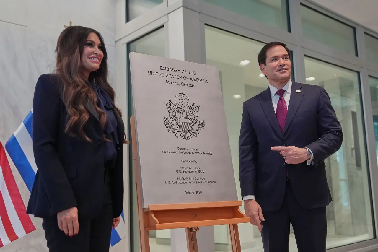 Two smiling officials beside a plaque reading Embassy of the United States, Athens, Greece