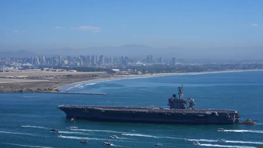 Aircraft carrier escorted by small boats into a bay, with San Diego skyline behind.