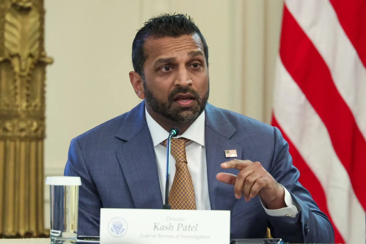 Man in blue suit speaking at table with name placard and American flag behind.
