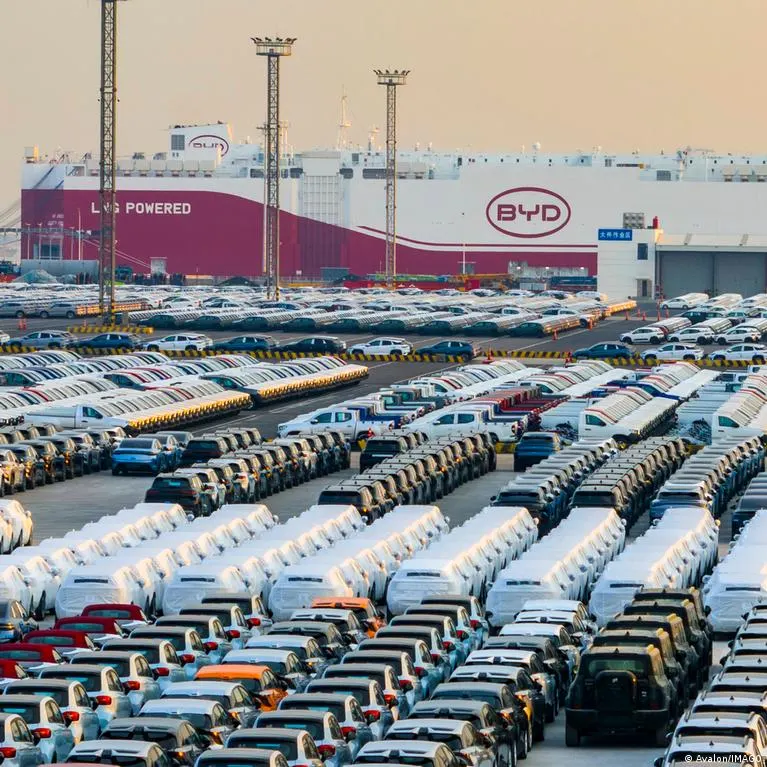 Rows of new cars parked at a port beside a BYD-branded car carrier ship.