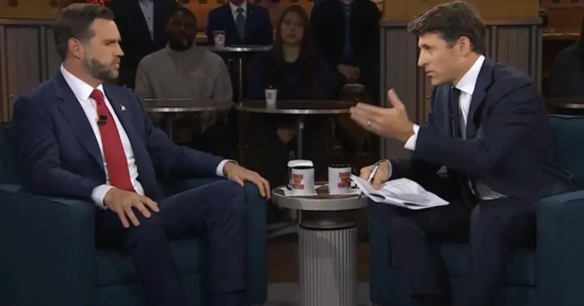 Two men seated in armchairs during a televised interview, with an audience in the background.