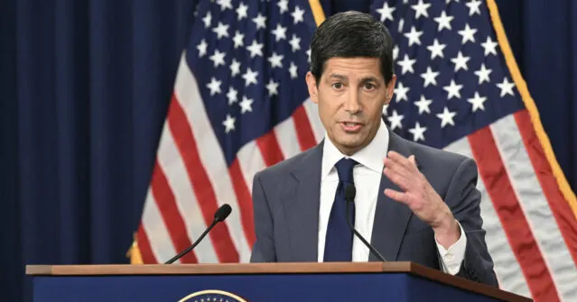 A man in a suit speaking at a podium with American flags behind him, gesturing with one hand.