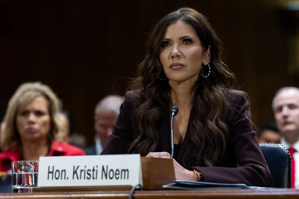 Woman in dark blazer seated at hearing table with nameplate and microphone, others behind her.