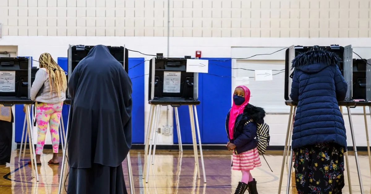 Voters at privacy booths in a gym, with a child in a pink hijab watching.