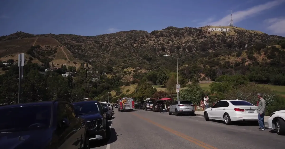 Parked cars line a road below the Hollywood Sign on a hillside, with a fire truck nearby.