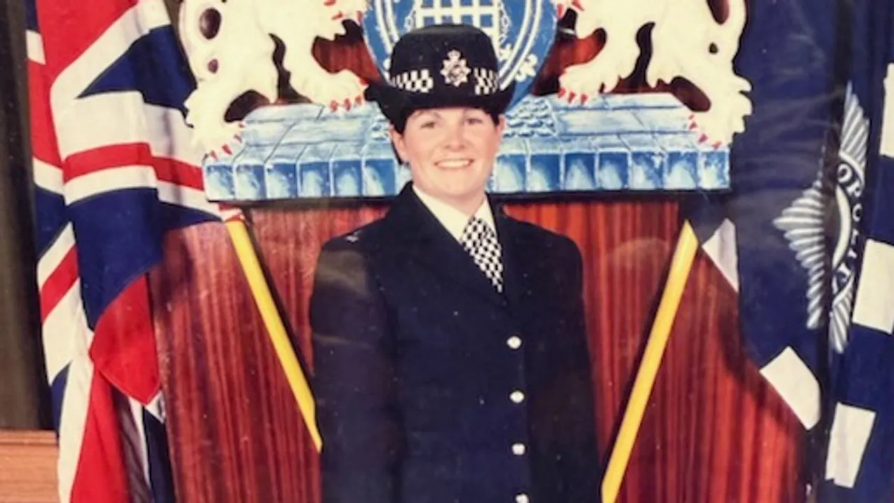 Smiling uniformed officer in peaked cap before a podium with the Metropolitan Police crest and flags.