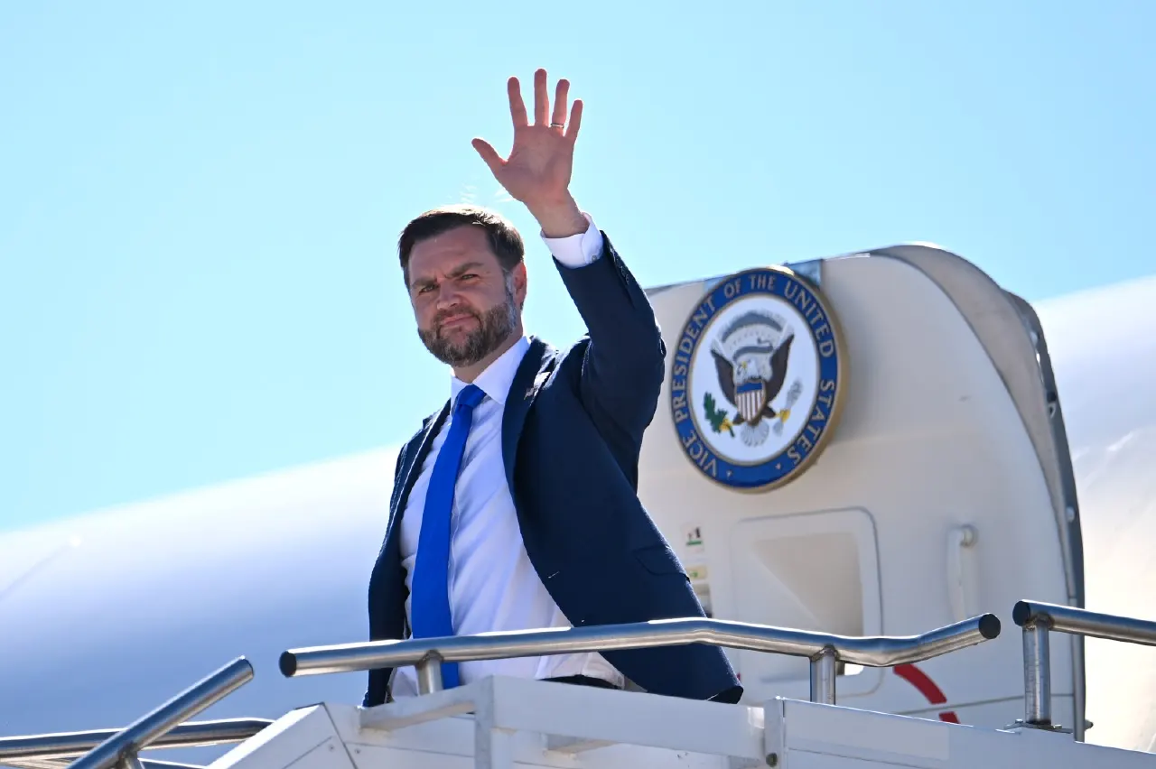 A man in a navy suit and blue tie waves from an aircraft stairway, with a Vice President seal visible.