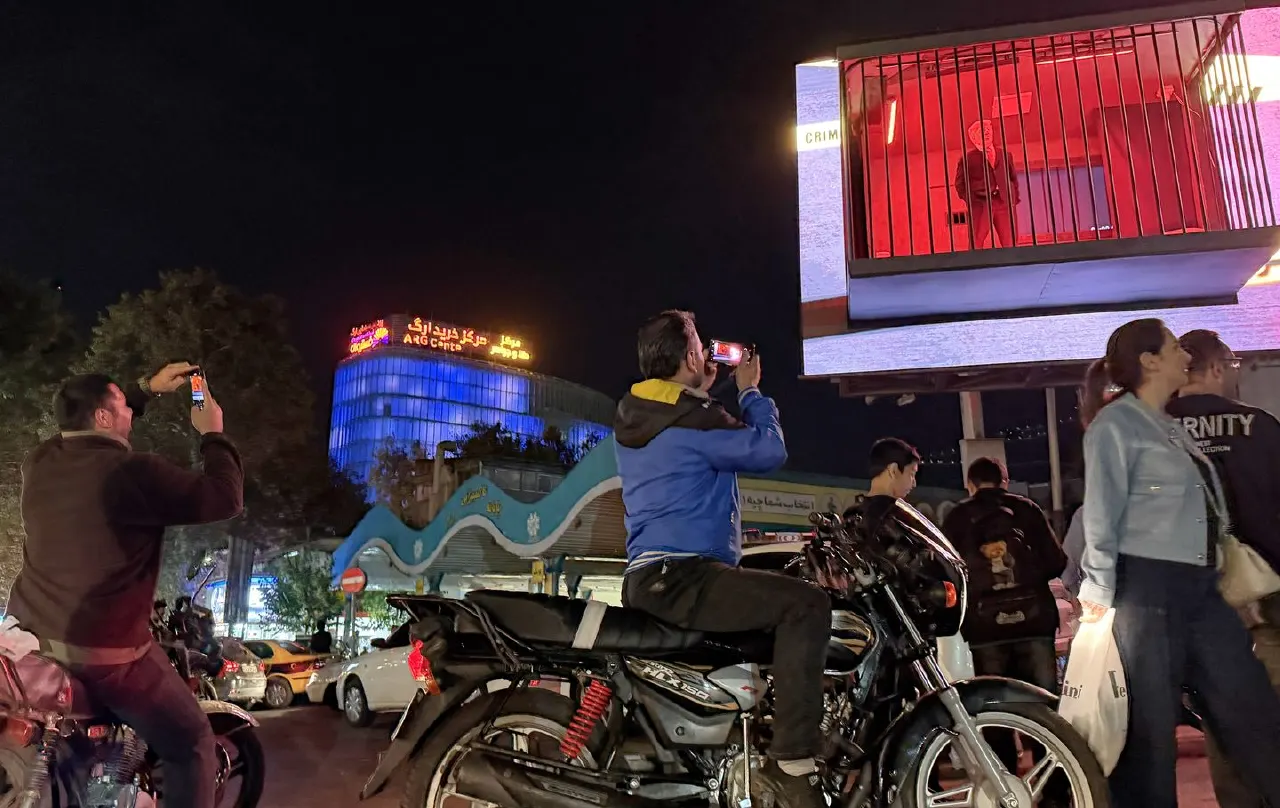 People photographing a lit city street at night, with motorcycles, cars and a illuminated building.