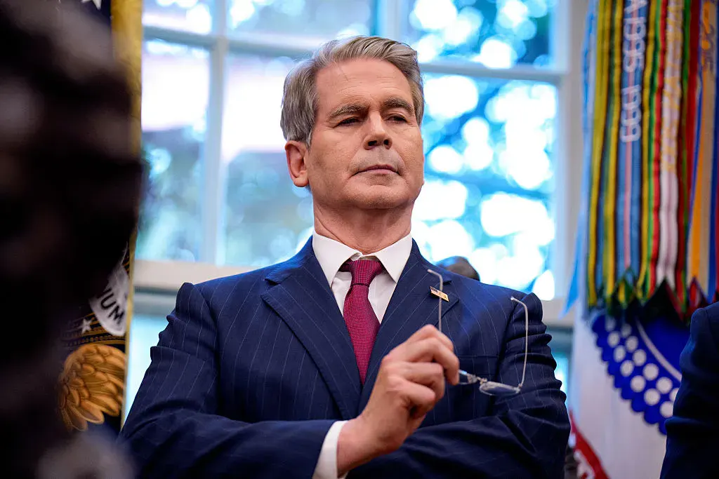 Man in navy pinstripe suit holding eyeglasses in a formal office with ribbons and window.
