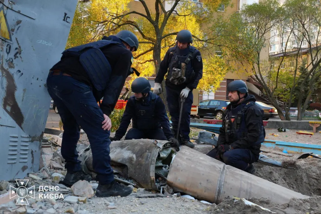 Rescue workers in helmets examine a large missile fragment in a Kyiv courtyard, with an agency watermark.
