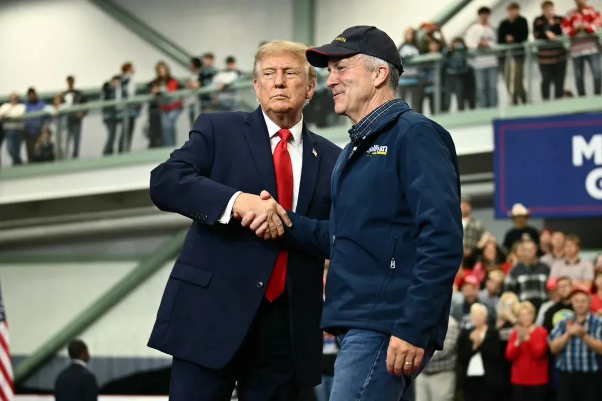 Two men in suits and jackets shake hands on stage before a crowd at an indoor event.