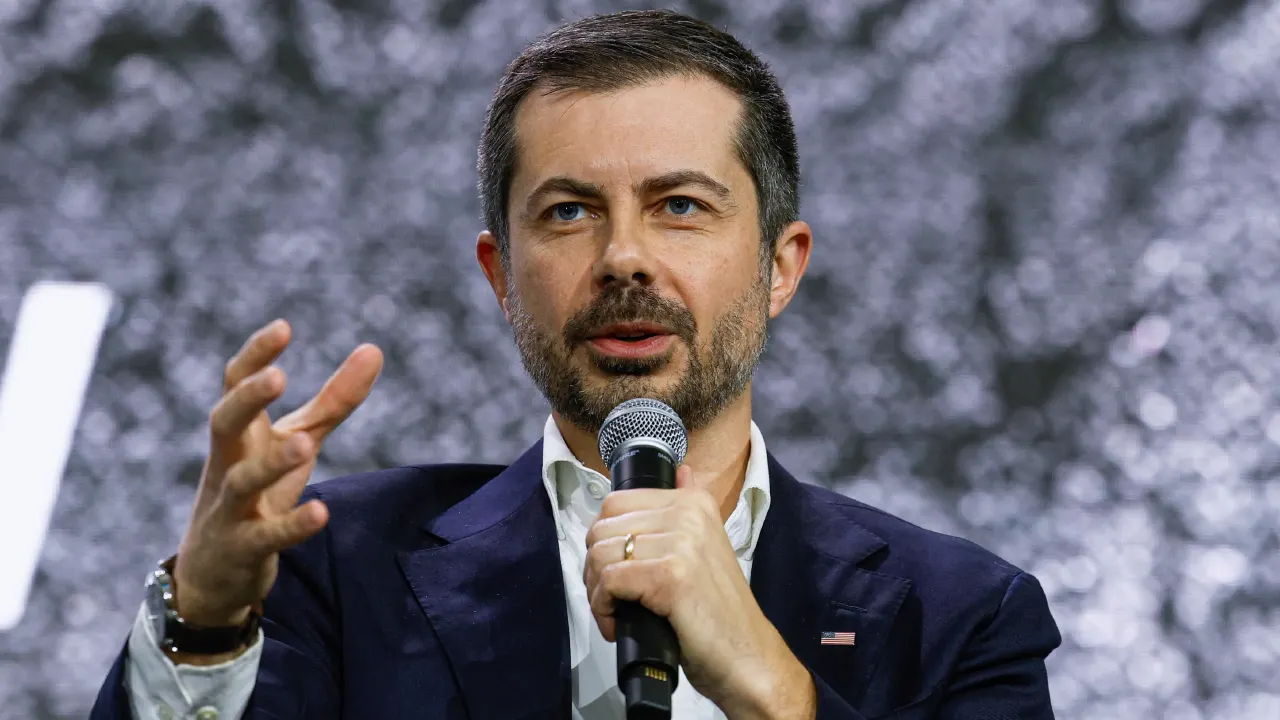 A man in a navy blazer speaks into a microphone, gesturing with one hand, against a sparkly backdrop.