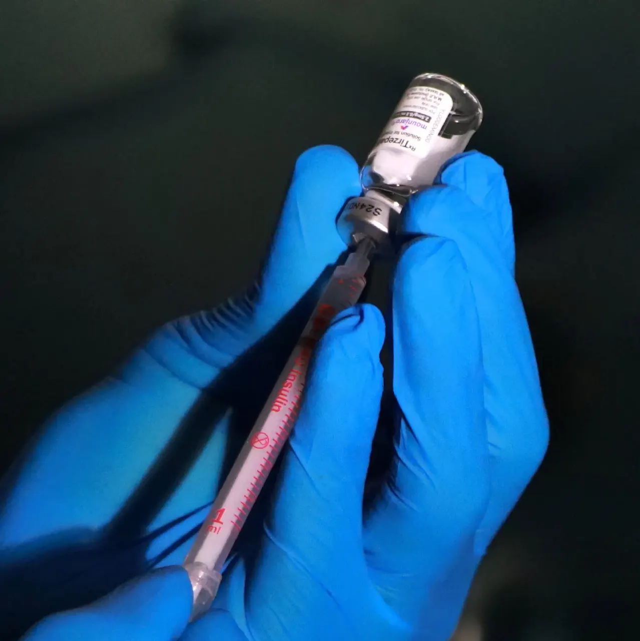 Gloved hands drawing liquid from a vial into a syringe against a dark background.