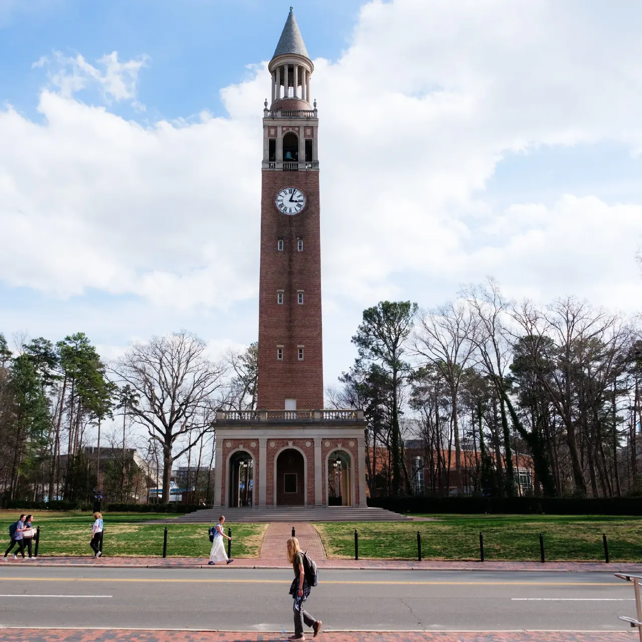 Brick clock tower on a campus lawn with students walking past, under cloudy sky.