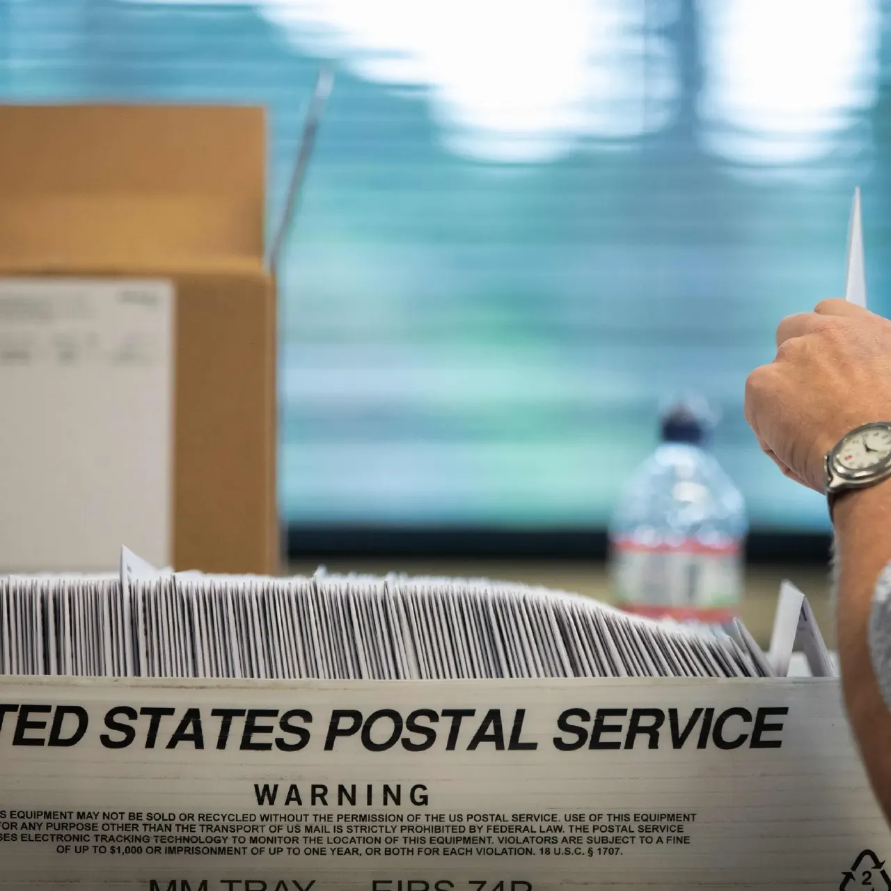 Hand holding a mail ballot over a United States Postal Service tray stacked with envelopes.