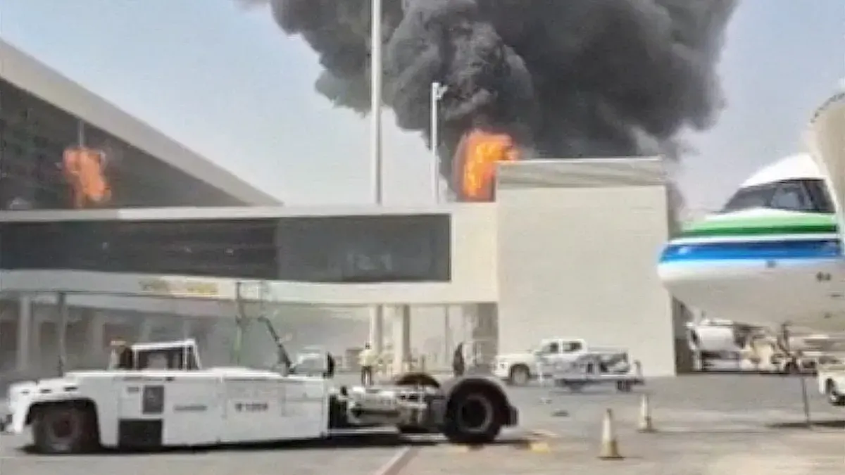 Large black smoke and flames rise behind an airport terminal, with an aircraft and vehicles nearby.