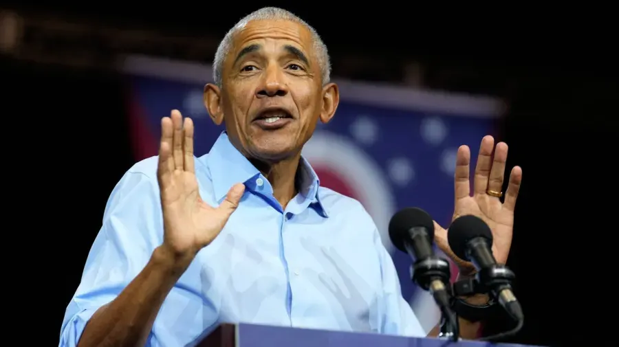 Older man in light blue shirt gesturing at a podium with microphones, speaking to an audience.