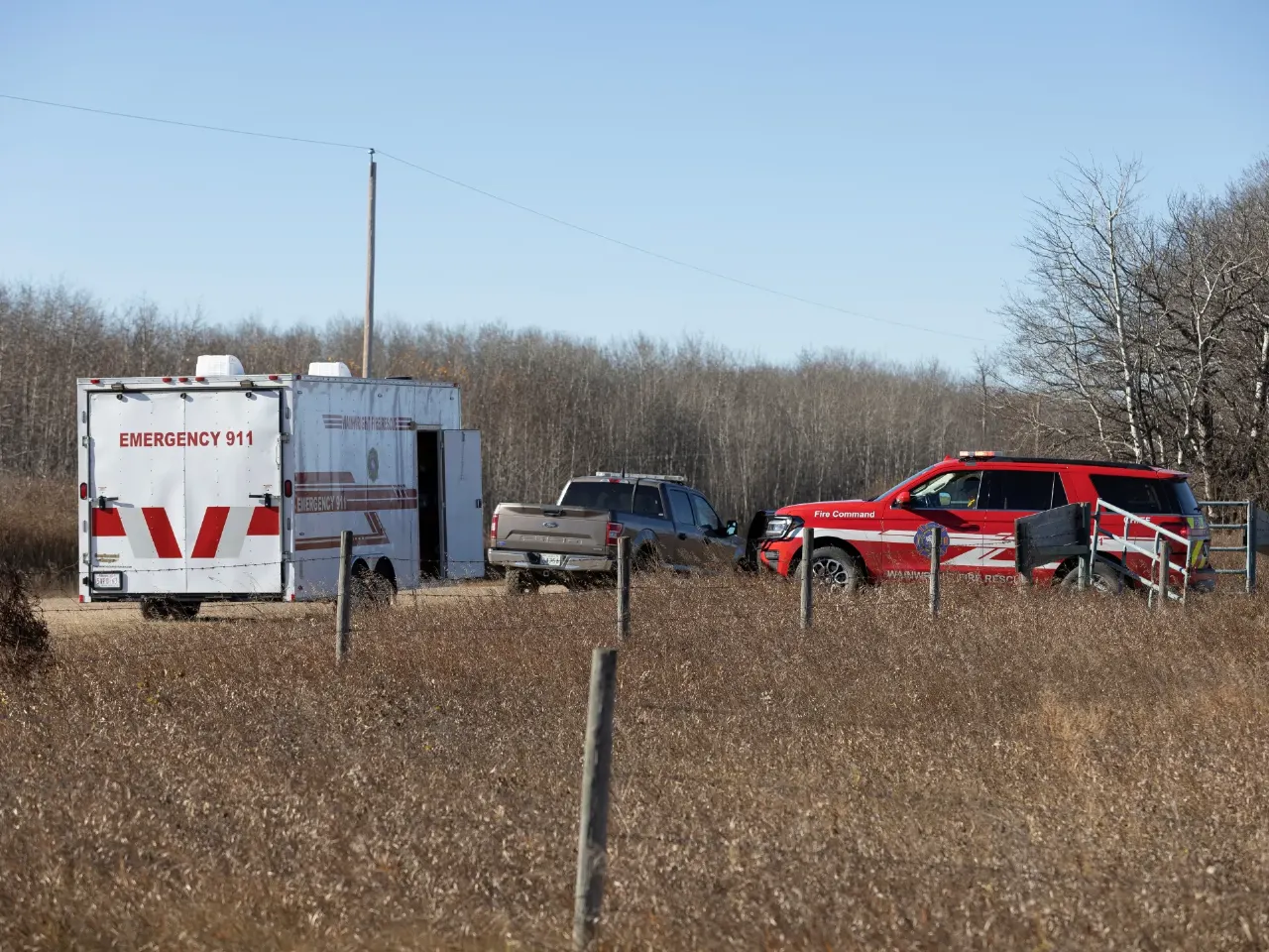 Emergency 911 trailer and red fire command vehicle parked in a dry field near woods.