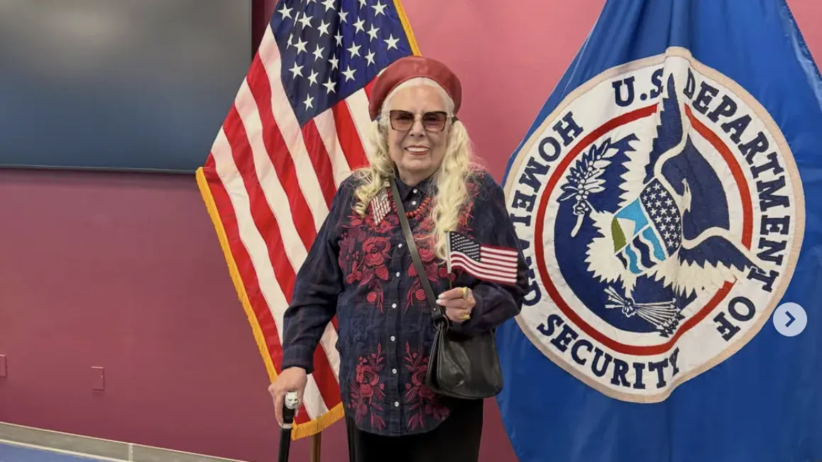 Smiling older woman holding small American flag beside U.S. flag and Department of Homeland Security seal.