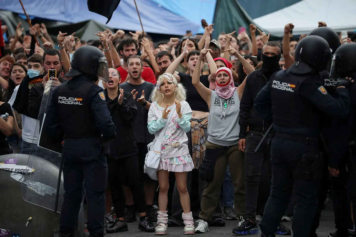 Crowd chanting and raising hands facing riot police in helmets with shields during a street protest.