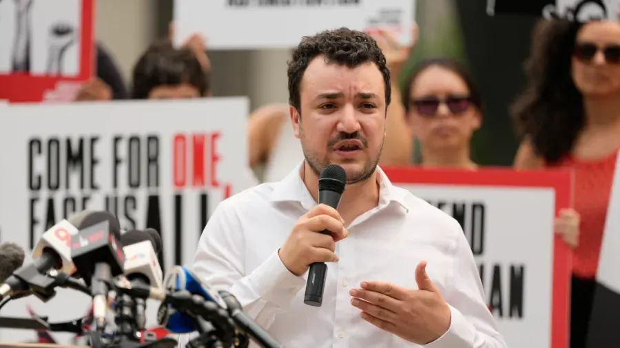 Man speaking into microphone at outdoor rally before protest signs reading "Come for one, face us all".