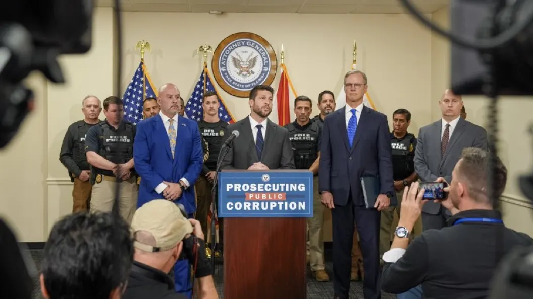 Officials at a podium with a 'Prosecuting Public Corruption' sign, flags and seal, with uniformed officers behind.