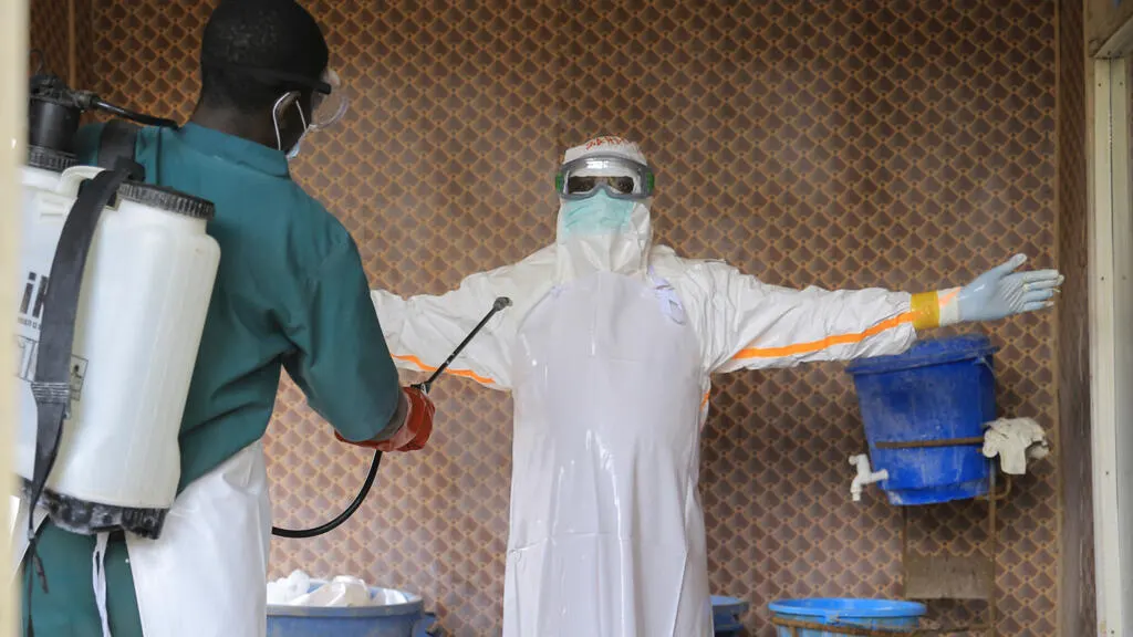 Worker in white protective suit and goggles arms outstretched while another sprays disinfectant nearby.