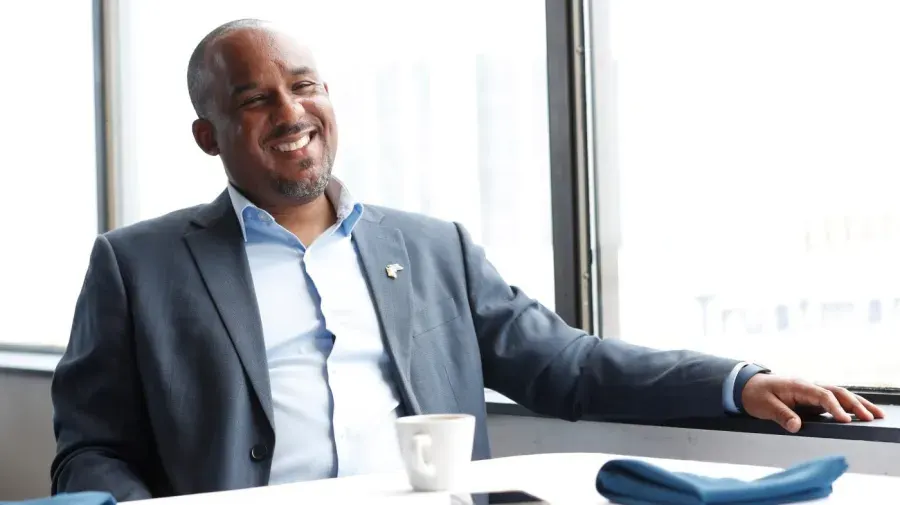 Smiling man in grey suit and blue shirt seated at table by window with coffee cup.