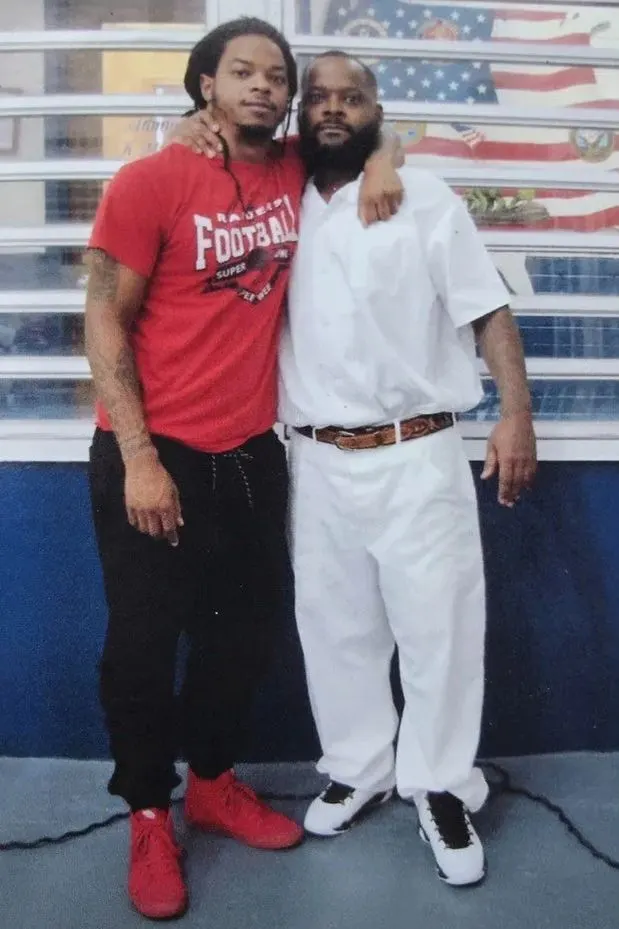 Two men posing together in front of a window with American flags, one in red shirt.