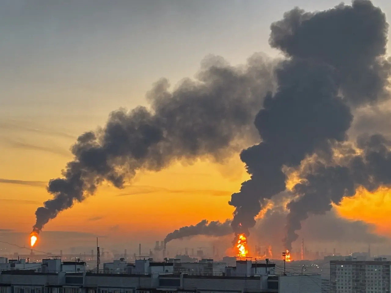 Thick black smoke and fire rising from industrial refinery facilities at sunset over a city
