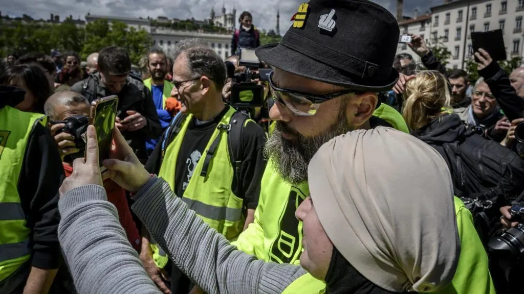 Crowd of people in yellow high-visibility vests at a street protest, one person filming with a phone.