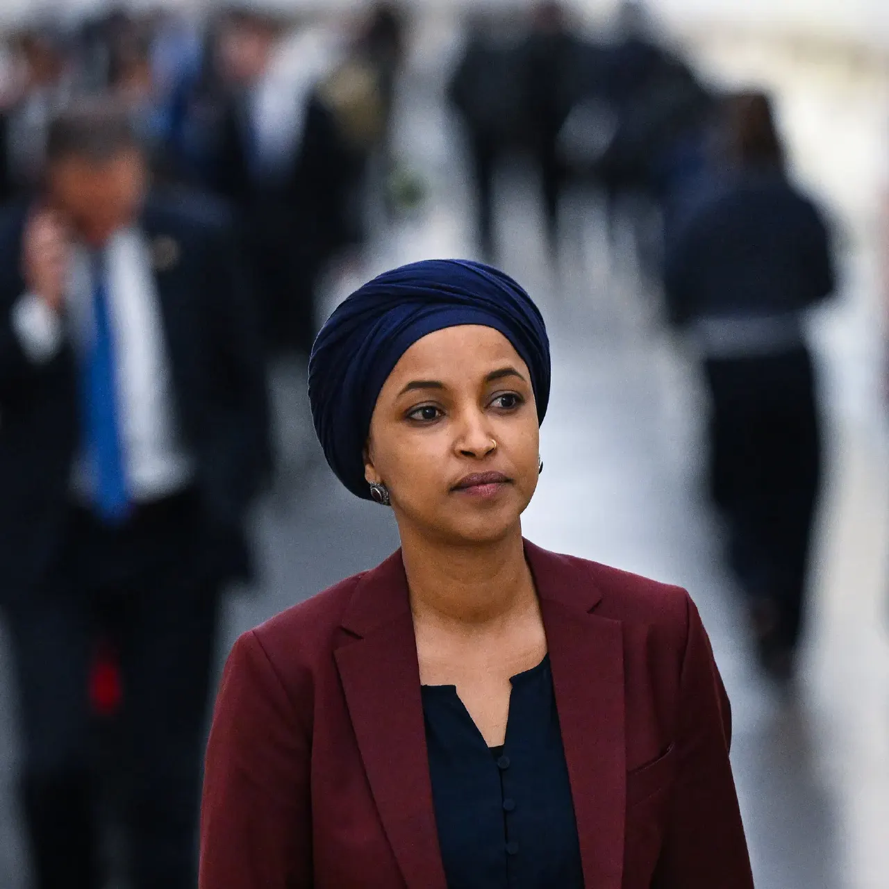 Woman in navy head wrap and maroon blazer walking through a busy blurred hallway