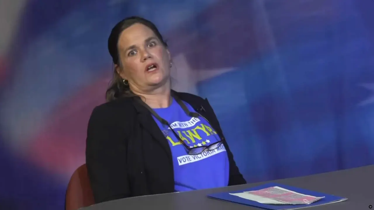 Woman in black blazer and blue campaign shirt seated at TV studio desk, mid-speech, eyes wide.
