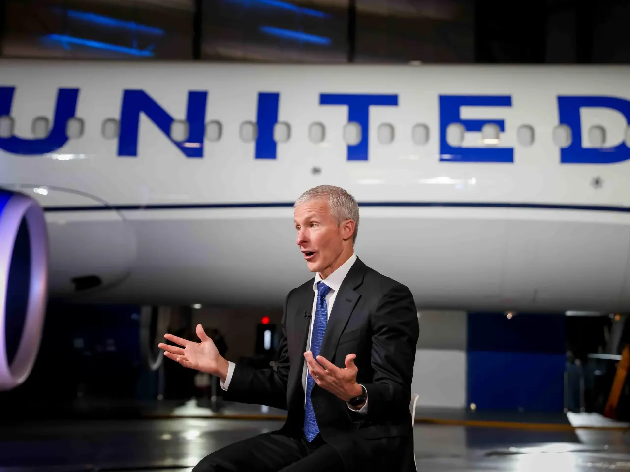 Man in dark suit gestures while speaking in front of a United Airlines jet.