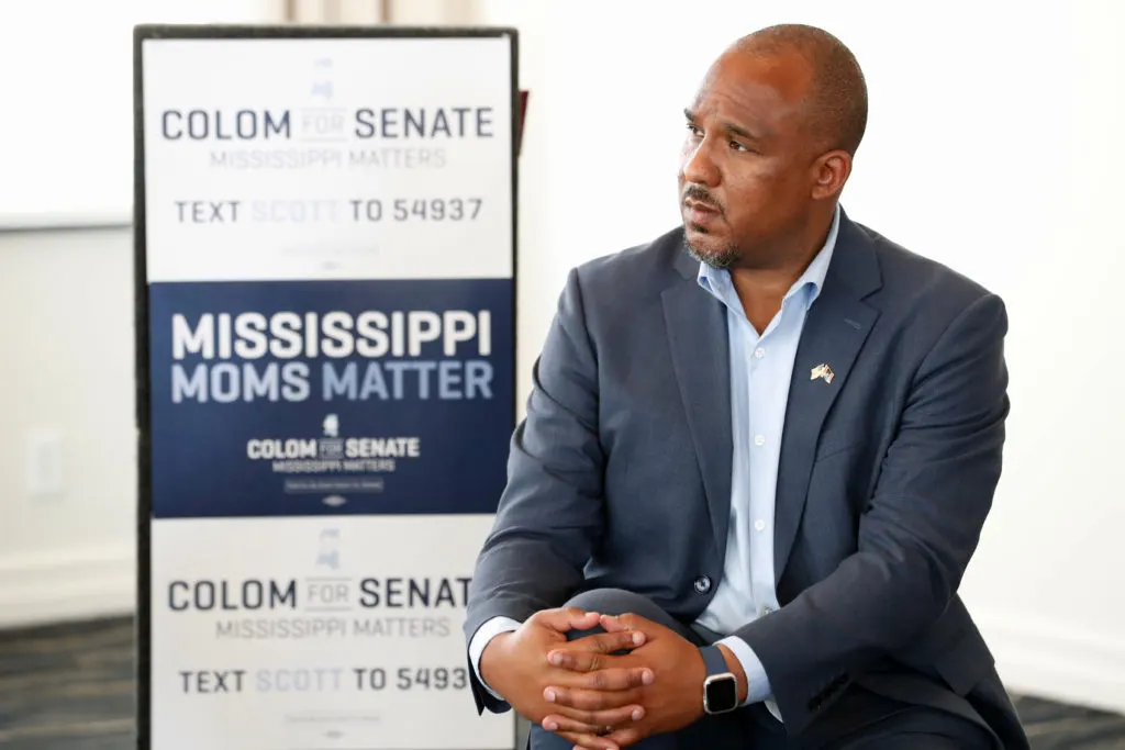 Man in suit seated beside Colom for Senate campaign sign reading Mississippi Moms Matter.