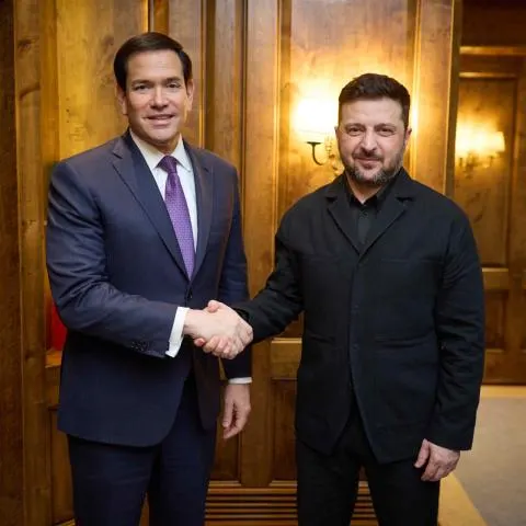Two men in suits smiling and shaking hands in front of a wood-paneled wall.