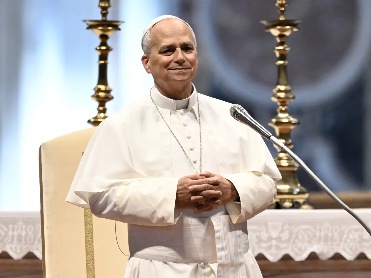 Man in white papal vestments and skullcap smiling, hands clasped, at a microphone before gold candlesticks.