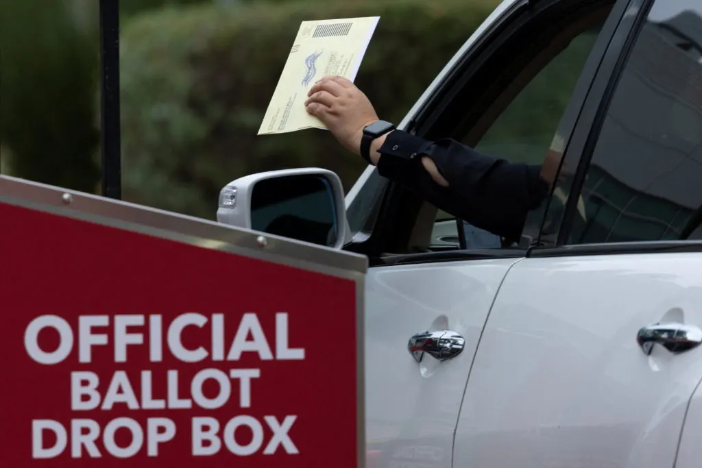 Arm from a car window holding a ballot envelope above an official ballot drop box.