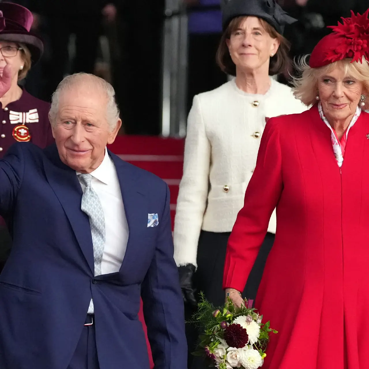 Older man in navy suit waves beside woman in red coat and hat, with a third woman behind.