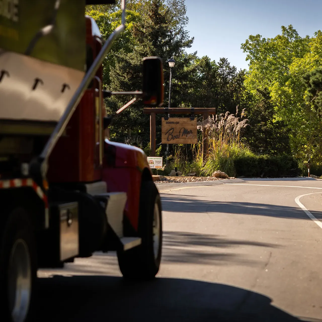 Red truck driving on road past Bow Mar town entrance sign among trees
