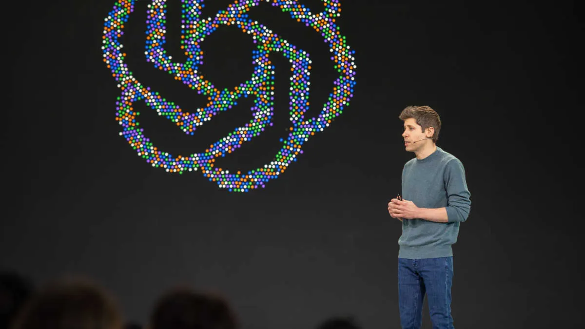 Man in grey sweater on stage beside large glowing multicolored swirl logo, audience silhouetted below.