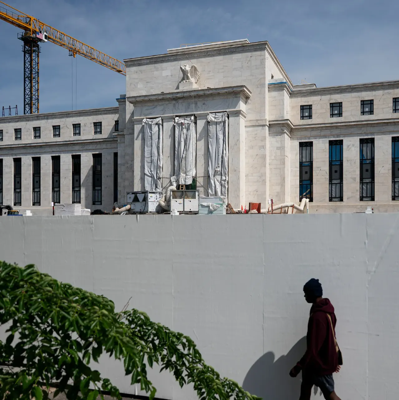 Federal Reserve building facade with eagle emblem, under renovation behind a white construction wall, pedestrian walking