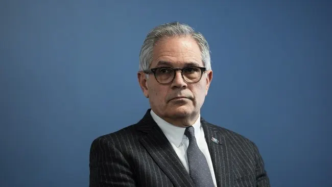 Portrait of a gray-haired man in glasses and pinstripe suit against blue background