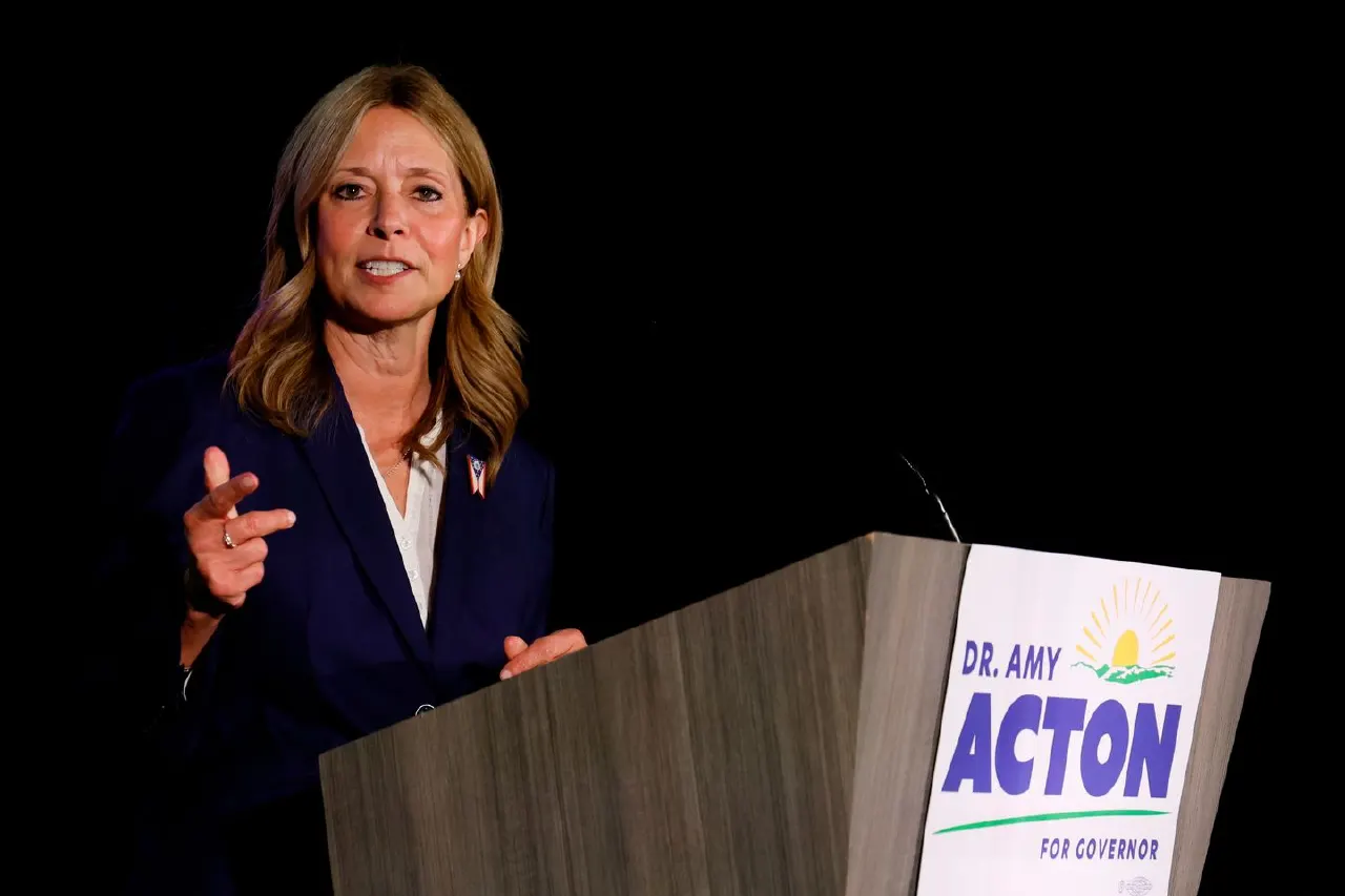 Woman in navy blazer speaking at podium with campaign sign reading Dr. Amy Acton for Governor
