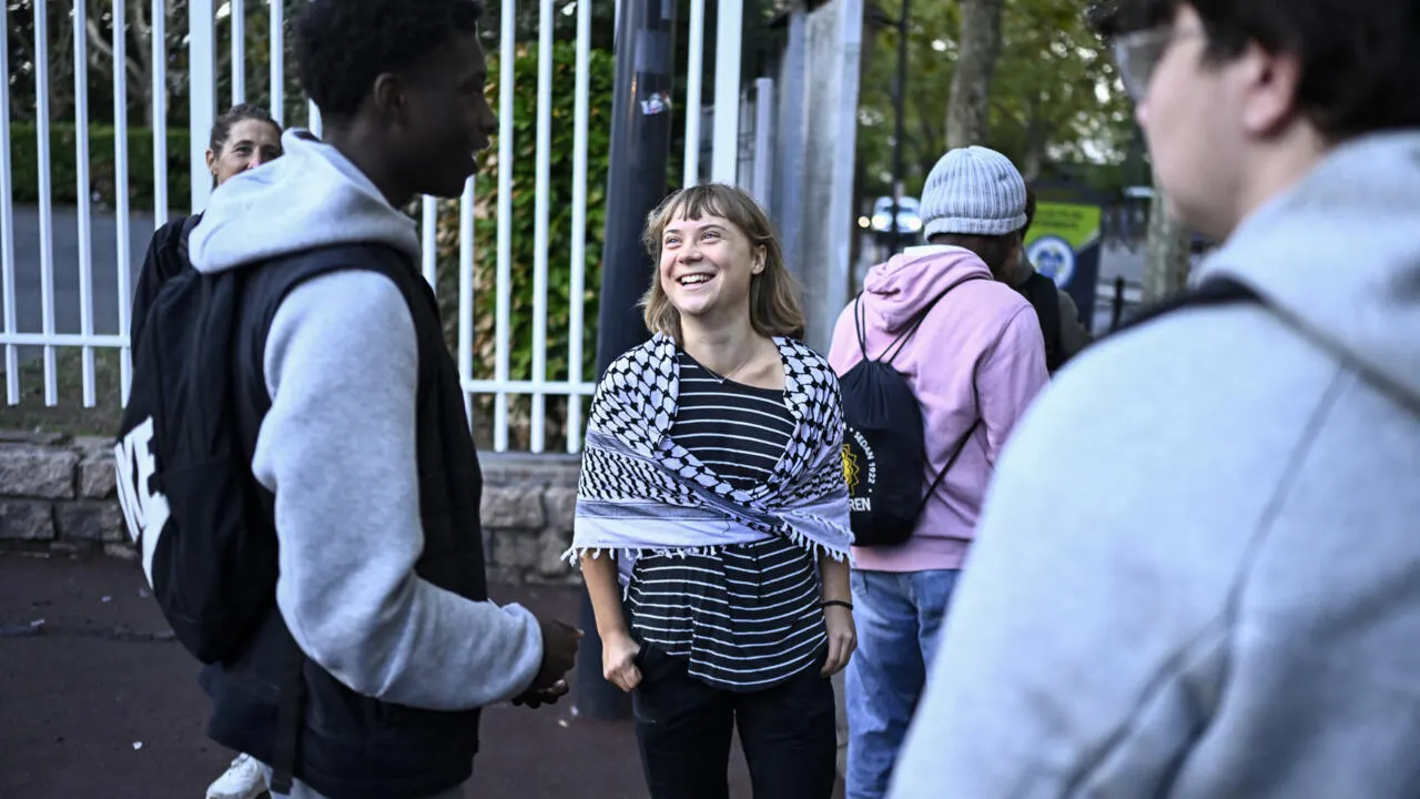 Smiling young woman in keffiyeh and striped shirt talks with students outside a gated school