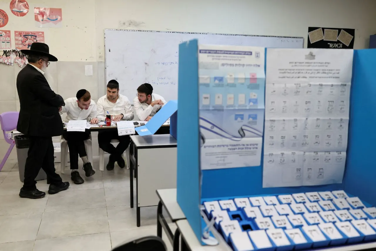 Men at a polling station table beside voting booths with ballot papers and sample ballots.
