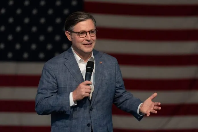 Man in glasses and blue suit holding microphone, gesturing, in front of American flag.