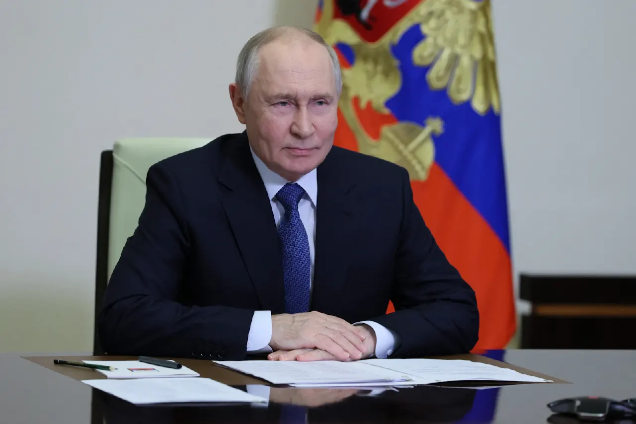 Man in dark suit and blue tie seated at desk before a Russian state flag with coat of arms.