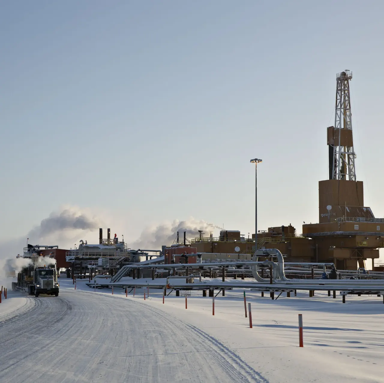 Snowy industrial gas facility with pipelines, drilling rig, and a truck on icy road