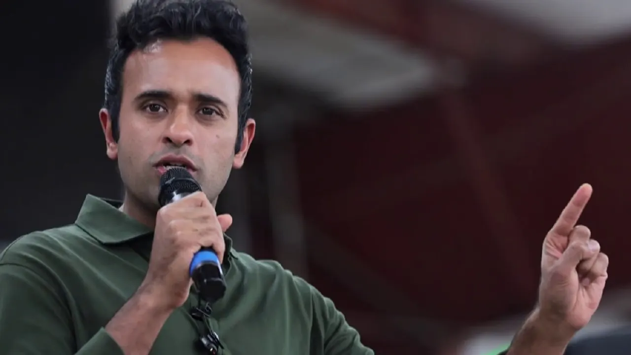 A man in a green polo speaks into a microphone while gesturing with one raised finger.